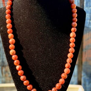 May include: A necklace featuring round, reddish-orange beads. The beads are strung together, creating a continuous loop. The necklace is displayed on a black velvet bust, with a blurred background of blue and white.