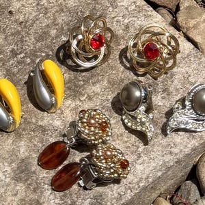Könnte beinhalten: Eine Sammlung verschiedener Vintage-Ohrringe. Dazu gehören goldfarbene Wirbelohrringe mit roten Zentren, silberne und gelbe gebogene Ohrringe, perlenartige Ohrringe und Perlenohrringe mit hängenden braunen Akzenten. Die Ohrringe liegen auf einer strukturierten Steinoberfläche.
