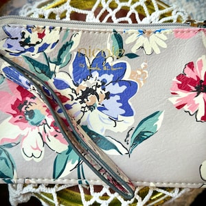 Op de afbeelding: Een polsportemonnee met bloemenprint in roze, blauw en crème. De portemonnee heeft een ritssluiting en een polsbandje. Het ontwerp bevat grote, gestileerde bloemen op een lichtgrijze achtergrond. De merknaam "nicole" is zichtbaar.