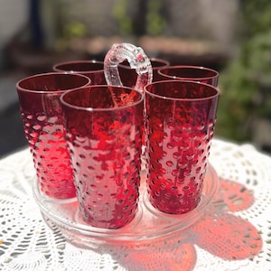 May include: A set of six deep red glass tumblers with a raised hobnail texture, arranged in a circular clear glass carrier. The carrier features a central clear glass handle with decorative detailing, providing a structured display for the glasses.