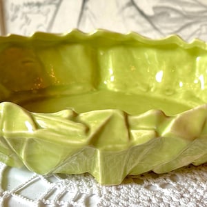 May include: A lime green ceramic bowl with a textured, ruffled exterior that mimics the appearance of layered leaves. The bowl has a glossy finish and a wide, shallow, oval-shaped opening with irregular, jagged edges along the entire rim.