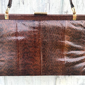 Peut inclure: Un sac à main rectangulaire doté d'une texture en peau de serpent marron foncé avec une finition brillante. Le sac comprend un cadre supérieur rigide avec des ferrures en métal doré, un fermoir central et des anses fines fixées aux coins.