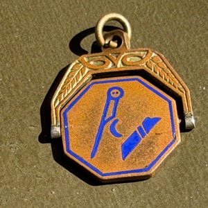 May include: A bronze-toned octagonal pendant featuring a central blue enamel design of a compass and a square. The pendant is suspended from a decorative metal bail with leaf-like engravings and includes a small circular loop at the top for hanging.