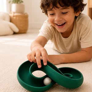 May include: A dark green, infinity-shaped marble run toy with a smooth, curved track design. A small, solid white sphere rests within the centre of the track, which features a continuous loop structure for rolling the ball along the surface.