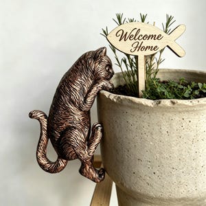 Op de afbeelding: Een bronskleurig kattenbeeldje met gestructureerde vacht dat aan de rand van een beige gespikkelde keramische bloempot hangt. In de pot staat een groene plant en een lichtgekleurde houten visvormige plantenmarker met de tekst Welcome Home.