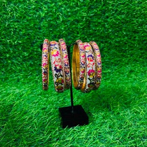 Op de afbeelding: Een set van zes ronde armbanden gepresenteerd op een zwarte standaard. Elke armband heeft een witte basis met een kleurrijk bloemenpatroon in roze, gele, blauwe en groene tinten, geaccentueerd door kleine metalen kralen langs de randen.