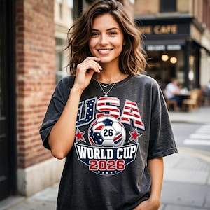 Pu&ograve; includere: T-shirt grigio scuro con grafica USA World Cup 2026. Il design presenta un pallone da calcio, stelle e la scritta "USA" in rosso e blu. La maglietta &egrave; in stile casual.