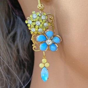 Può includere: Orecchini dorati con accenti di fiori blu e gialli. Gli orecchini presentano un gruppo di piccoli strass e una pietra blu a forma di lacrima pendente.