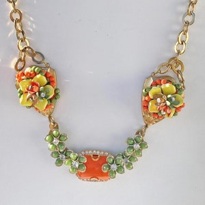 Puede incluir: Un collar de cadena dorada con tres colgantes en forma de flor naranja, verde y amarilla. El colgante central es una piedra rectangular naranja con diamantes de imitación alrededor de los bordes.