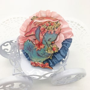 Op de afbeelding: Een vintage Paasdecoratie met een blauwe vogel met een muts en paraplu, omringd door roze en blauwe stof. De vogel houdt bloemen vast en op de paraplu staat "An Easter Time Hello". De decoratie staat in een witte koets.