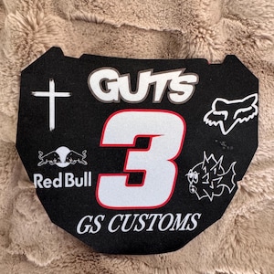 May include: Black motocross number plate with the number 3 in white and red. The plate features the text "GUTS" and "GS CUSTOMS", along with logos for Red Bull and Fox Racing. A white cross and a spiky graphic are also visible.