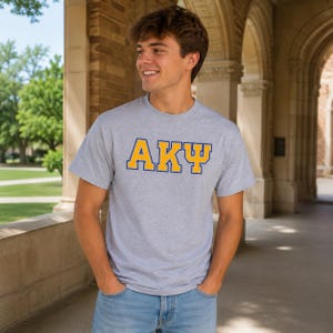 May include: A light gray t-shirt with the letters "AKΨ" in gold with a blue outline. The person is wearing blue jeans and standing outside in front of a brick building.