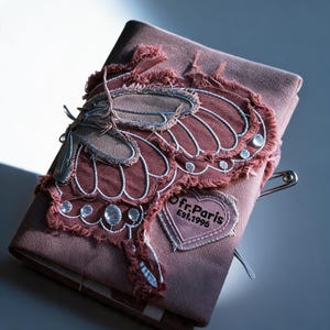 以下が含まれることがあります： 縁がほつれた大きな刺繍の蝶と銀色のステッチが施された、ダスティローズ色の布製ノートカバー。右下のハート型のパッチには「Ofr.Paris Est.1996」の文字が記されている。側面には銀色の安全ピンが取り付けられている。