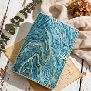 May include: A rectangular notebook cover featuring a textured blue and teal marbled pattern with gold-colored vein accents. The cover has a single small white button closure on the right side, securing the front flap against the main body.