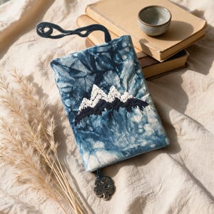 May include: A rectangular fabric book cover featuring a blue and white tie-dye pattern with an embroidered mountain range in white and dark blue thread. The cover includes a dark blue curved bookmark attachment and a silver-toned tree charm.