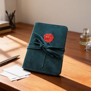 Puede incluir: Un cuaderno de pana de color verde azulado oscuro con textura acanalada vertical y un cierre de lazo de tela a juego. Una rosa roja bordada está centrada en la portada, situada justo encima del cordón que envuelve el cuaderno.