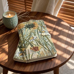 May include: A rectangular fabric journal cover featuring a woven botanical pattern of green, yellow, and white wildflowers. The cover includes a cream-coloured elastic cord closure secured by a single round button on the front edge.