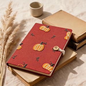 May include: A rectangular notebook with a textured dark red cover featuring embroidered orange and white pumpkin motifs. The notebook is secured with a cream-coloured braided cord and a single wooden button closure on the right side edge.