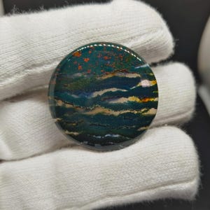 Puede incluir: Un cabujón de piedra preciosa circular y pulido que presenta una base de color verde bosque profundo con bandas horizontales onduladas de azul verdoso oscuro, crema y sutiles inclusiones de color rojo anaranjado. La superficie es lisa y brillante.