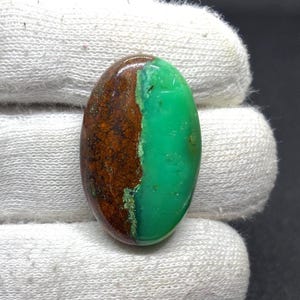 May include: An oval-shaped cabochon gemstone featuring a distinct split color pattern. One side is a smooth, vibrant apple green, while the other side is a textured, dark brown matrix with earthy tones. The stone has a polished, glossy finish.