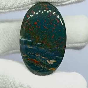 Puede incluir: Un cabochon de piedra de sangre de forma ovalada con una base de color verde bosque profundo, que presenta puntos rojos brillantes distintos y dispersos, junto con sutiles bandas horizontales de inclusiones minerales blancas y marrones.