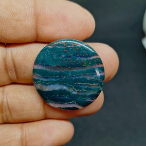 Puede incluir: Un cabujón de piedra preciosa circular y pulido que presenta bandas horizontales de color verde azulado profundo y verde oscuro con sutiles capas translúcidas y pequeñas inclusiones de color marrón rojizo en la superficie lisa.