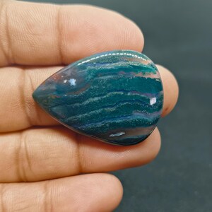 Puede incluir: Una piedra preciosa pulida en forma de lágrima con bandas horizontales en azul verdoso oscuro y verde bosque, áreas translúcidas e inclusiones sutiles. La piedra tiene una superficie lisa y reflectante con un borde redondeado.