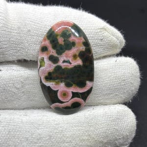 May include: An oval-shaped cabochon gemstone featuring a polished surface with a distinct pattern of dark forest green and soft pink circular inclusions. The stone displays a smooth, reflective finish with organic, mottled color variations throughout.