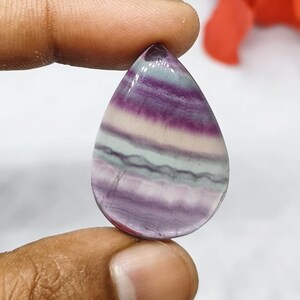 Può includere: Una pietra preziosa lucidata a forma di goccia con bande orizzontali di colori viola, bianco e verde pallido traslucidi. La pietra presenta una superficie liscia e lucida con una distinta stratificazione di colori in tutta la struttura.
