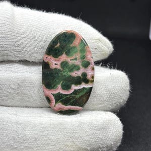 May include: An oval-shaped cabochon gemstone featuring a polished surface with a distinct pattern of deep forest green and soft pink swirling inclusions. The stone displays a smooth, rounded top and a glossy finish, showcasing natural color variations.