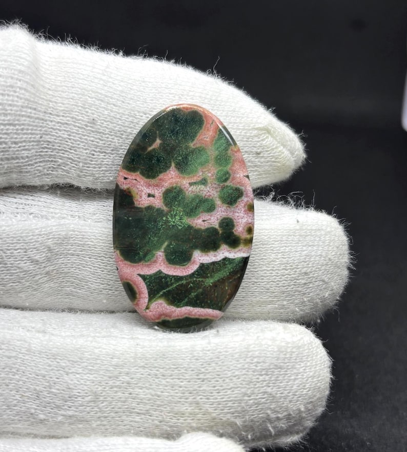May include: An oval-shaped cabochon gemstone featuring a polished surface with a distinct pattern of deep forest green and soft pink swirling inclusions. The stone displays a smooth, rounded top and a glossy finish, showcasing natural color variations.