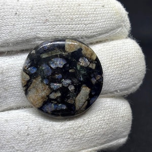 May include: A circular cabochon gemstone featuring a polished surface with a dark black base color. The stone displays a mosaic-like pattern of embedded fragments in shades of tan, beige, and iridescent blue, creating a high-contrast appearance.