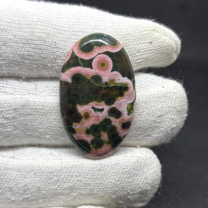 May include: An oval-shaped cabochon gemstone featuring a polished surface with a distinct pattern of dark forest green and deep olive circular spots contrasted against swirling bands of soft pastel pink and creamy white mineral inclusions.