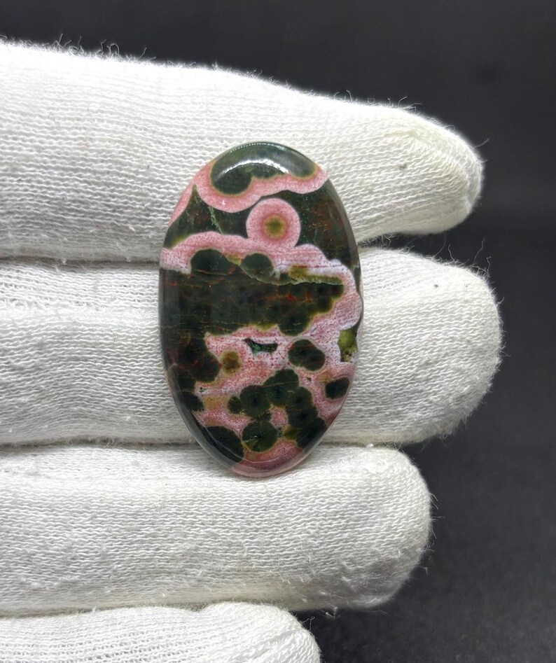 May include: An oval-shaped cabochon gemstone featuring a polished surface with a distinct pattern of dark forest green and deep olive circular spots contrasted against swirling bands of soft pastel pink and creamy white mineral inclusions.