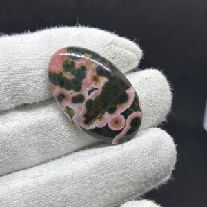 May include: An oval-shaped polished gemstone cabochon featuring a distinct pattern of deep forest green and dark brown patches contrasted by swirling bands and circular inclusions of soft pink and light rose colors throughout the smooth surface.
