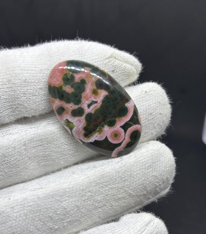 May include: An oval-shaped polished gemstone cabochon featuring a distinct pattern of deep forest green and dark brown patches contrasted by swirling bands and circular inclusions of soft pink and light rose colors throughout the smooth surface.