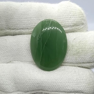 Könnte beinhalten: Ein ovaler, grüner Edelstein-Cabochon mit einer glatten, polierten Oberfläche, die durch feine weiße Aderungen gekennzeichnet ist. Der Edelstein zeigt eine tiefe, satte grüne Farbe mit natürlich vorkommenden Einschlüssen auf der Oberfläche.