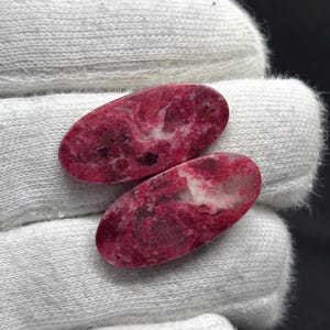 May include: Two oval-shaped cabochons featuring a vibrant magenta and deep pink color palette with marbled white and dark inclusions. The stones have a smooth, polished surface and are displayed against a textured white fabric background.