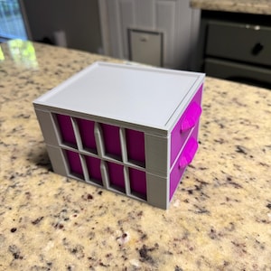 May include: A small, square storage box with a white top, gray frame, and magenta interior. The box has a grid-like design on the sides and two magenta tabs. The box is sitting on a speckled countertop.