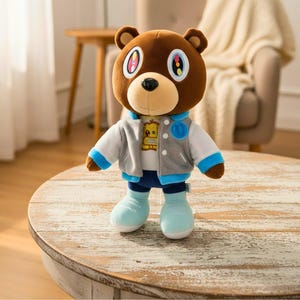 Puede incluir: Un oso de peluche marrón que lleva una chaqueta gris y blanca con detalles azules, una camisa blanca, pantalones azules y botas azul claro. El oso tiene ojos grandes con iris coloridos y una cara redonda y bronceada. Un parche en la chaqueta presenta un robot.