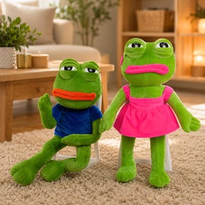 Op de afbeelding: Twee groene kikkerknuffels. De linker kikker draagt een blauw t-shirt en heeft oranje lippen. De rechter kikker draagt een felroze jurk en heeft roze lippen. Beide hebben grote, halfgesloten ogen en zitten op kleine witte stoeltjes.