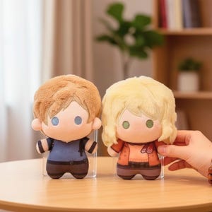 May include: Two plush dolls with large heads and small bodies. One doll has brown hair and wears a blue shirt and brown trousers. The other doll has blonde hair and wears an orange jacket and brown trousers. Both dolls have large eyes.