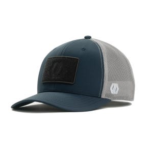 May include: A navy blue baseball cap with a gray mesh back. The cap features a black square patch on the front with a white logo. The brim is curved, and the cap has a button on top.
