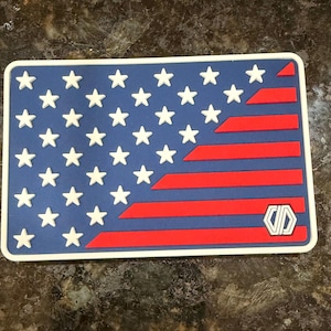 May include: A rectangular patch featuring a stylized American flag design with a blue field, white stars, and red stripes. The design includes a white border and a small white geometric logo in the lower right corner on a red stripe.