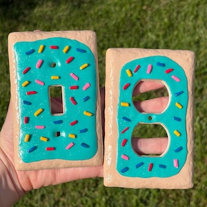 Op de afbeelding: Twee rechthoekige afdekplaatjes voor lichtschakelaars en stopcontacten in de vorm van gebakjes. Elk plaatje heeft een beige, getextureerde basis met een glanzende, turquoise glazuurlaag en verspreide, veelkleurige spikkels in rood, geel, roze en blauw.