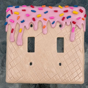 Op de afbeelding: Een dubbele lichtschakelaar wandplaat met een getextureerde beige wafelpatroon basis en een dikke laag roze glazuur die van de bovenrand afdruipt. Het glazuur is versierd met kleine, kleurrijke hagelslag in blauw, geel, rood en roze tinten.