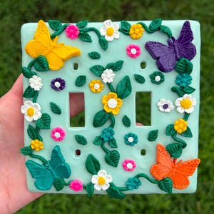 May include: A light mint green double light switch cover plate decorated with raised, colorful floral and butterfly accents. The design features yellow, purple, teal, and orange butterflies, along with small white, yellow, and pink flowers.