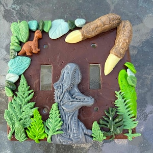 May include: A brown double light switch plate featuring a sculpted grey dinosaur head in the centre, a small orange dinosaur figure, two large brown dinosaur claws with cream-coloured nails, and various green and light blue sculpted leaves.