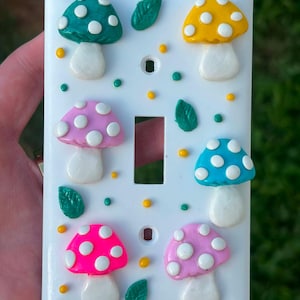 May include: A white rectangular light switch wall plate decorated with six three-dimensional mushrooms in teal, yellow, pink, blue, neon pink, and light pink, each featuring white spots, interspersed with small green leaves and yellow dots.