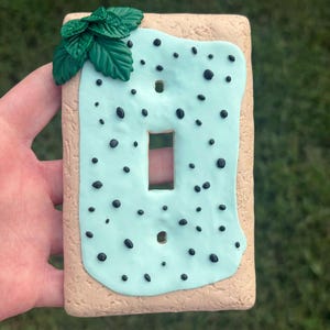 May include: A rectangular light switch cover plate designed to resemble a pastry. The base is tan with a textured surface, topped with a light blue icing layer featuring small black dots and a cluster of three green leaves in the top corner.